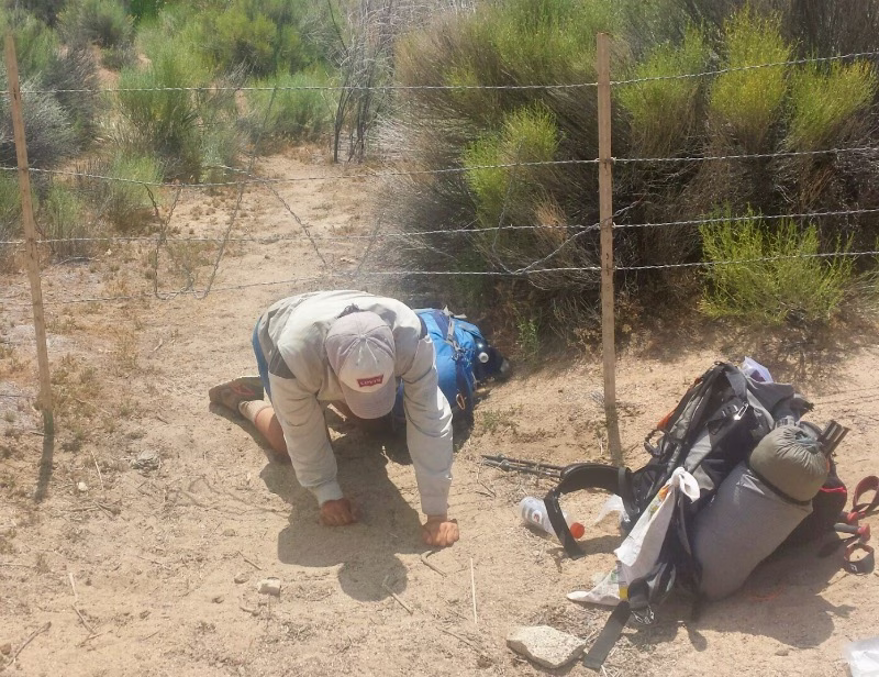 Arthur crawling under a barbed wire fence to access a public water source while backpacking in Southern California.